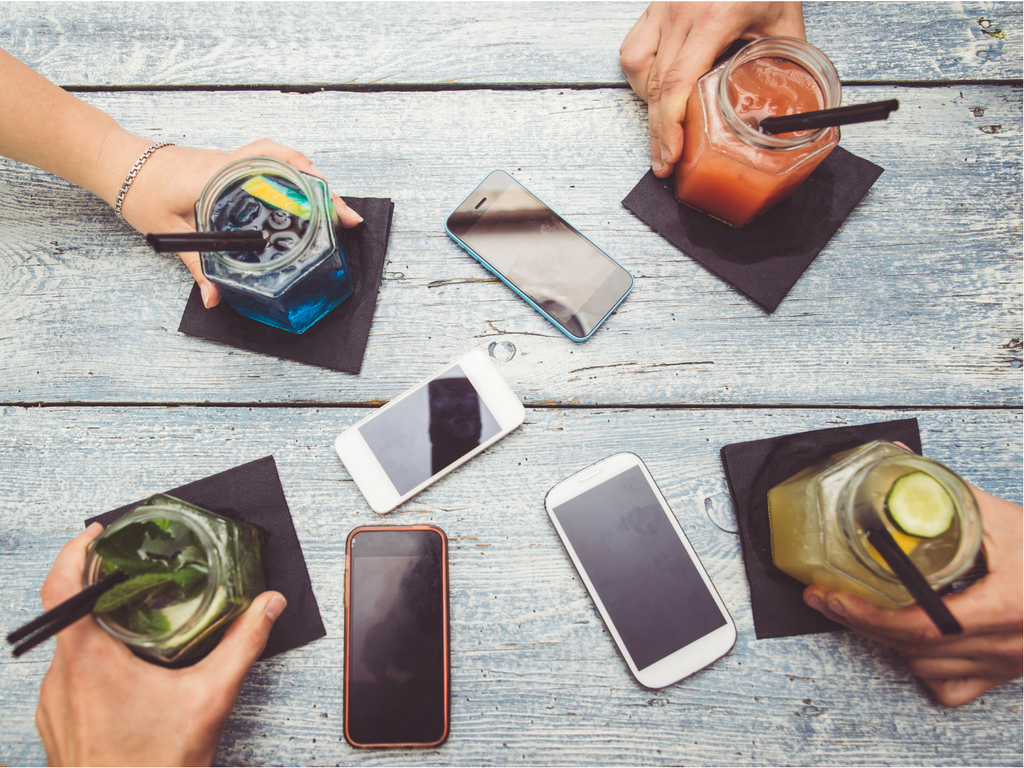 A spread of bright colored cocktails are held by hands, 4 phones screen up on the table.