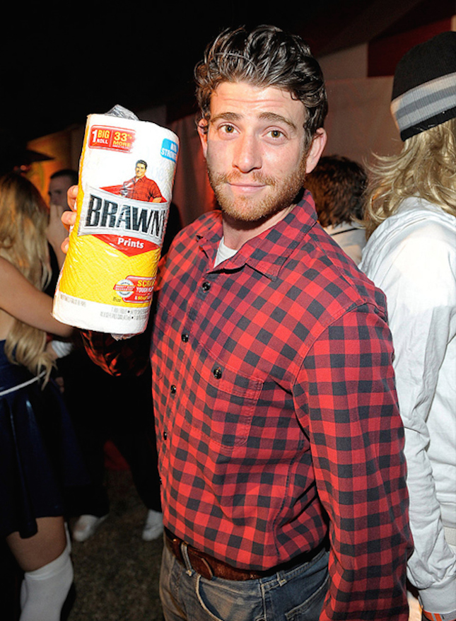 A man in a red and black plaid shirt holds a roll of Brawny paper towels