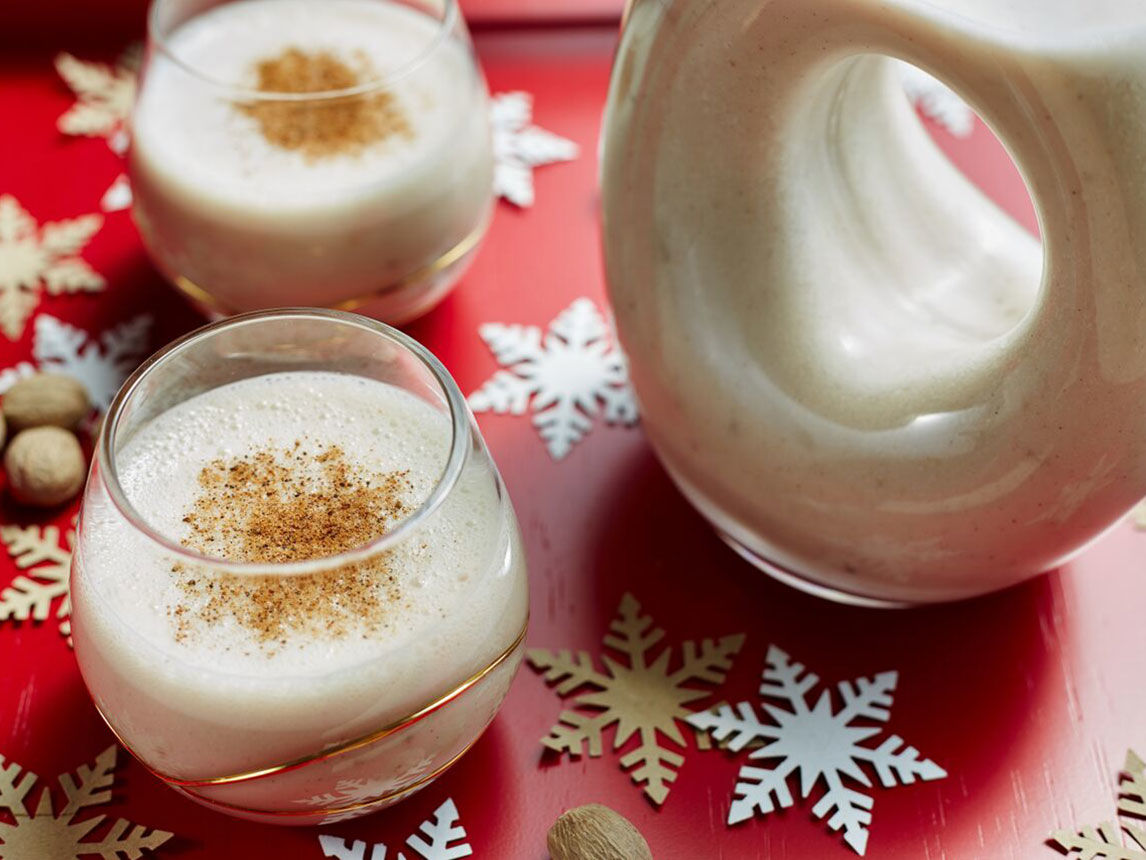 2 full glasses of eggnog sit on a table surrounded by paper snowflakes with a fun circular pitcher