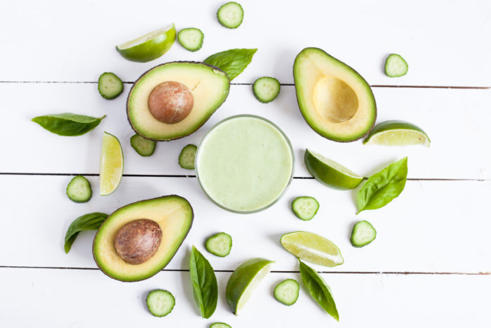 a green smoothie sits on a white table surrounded by halved avocados and basil leaves