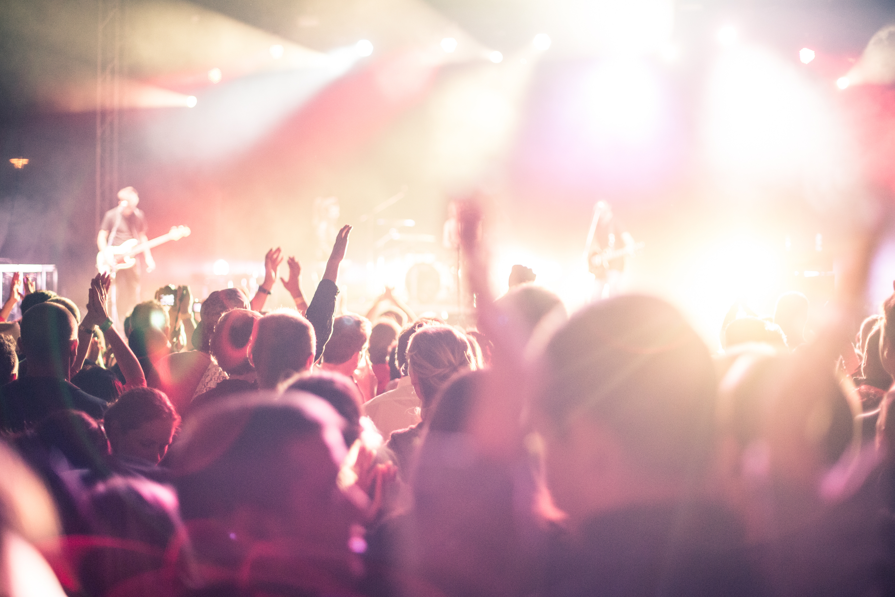 Fans cheer in a crowded space. A band sings on stage under bright lights.