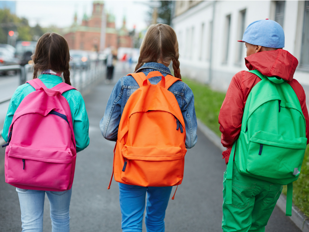 3 children walk to school wearing backpacks in pink, orange, and green