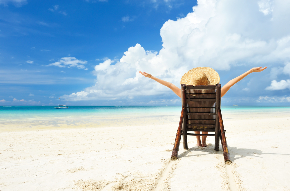 Woman on sunny beach wearing sunhat spreads arms wide