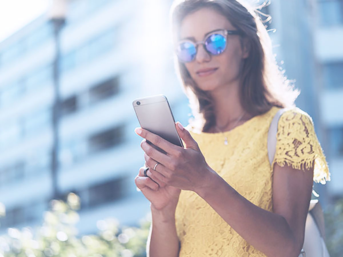A woman wears sunglasses and looks at her phone outside for Ibotta savings