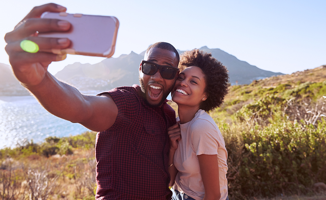 Two people smile outdoors with a mountain in the background. They're taking a selfie picture with a smart phone