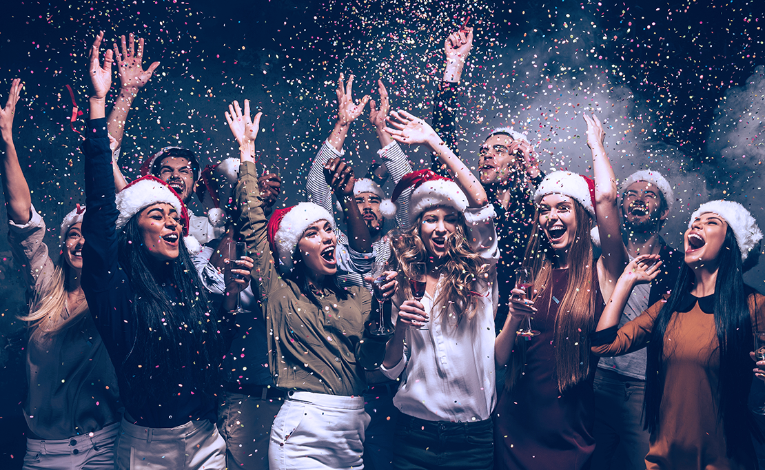 A group of eight friends wear santa hats and jump into white confetti, celebrating