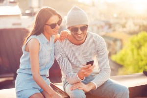 Two people wearing sunglasses sit outside and look at a single smart phone.