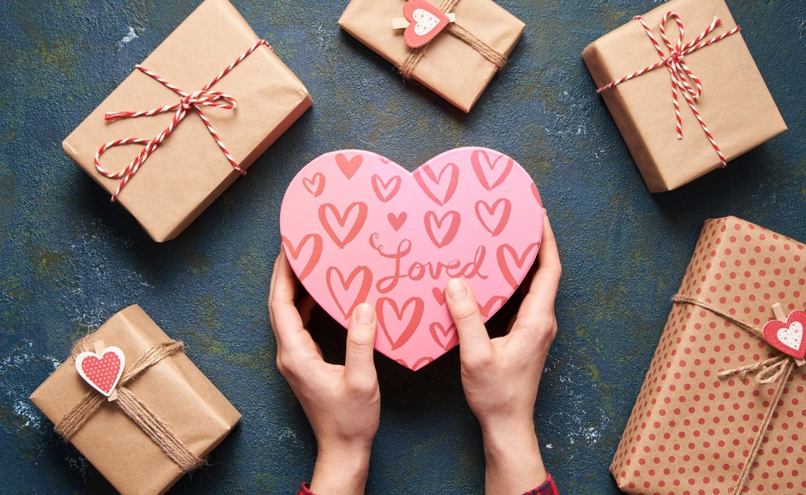 A bunch of gifts wrapped in brown paper surround a heart box held by hands