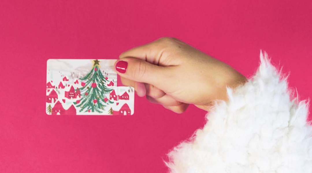 A woman's hand leading from an arm with a white fuzzy coat holds a gift card with a Christmas tree on it