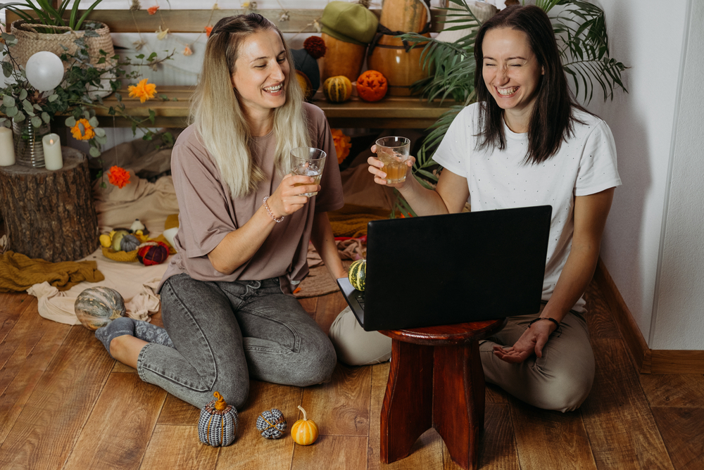 Two women sit on the floor, drinking white wine. One has a laptop on their lap.