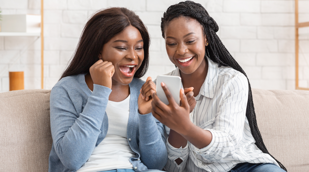 Two women sit on a couch and look at one smartphone screen together