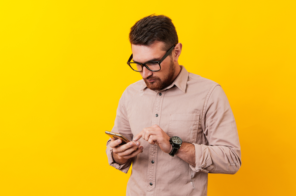 A man with glasses looks down at the smartphone in his hand in front of a yellow background