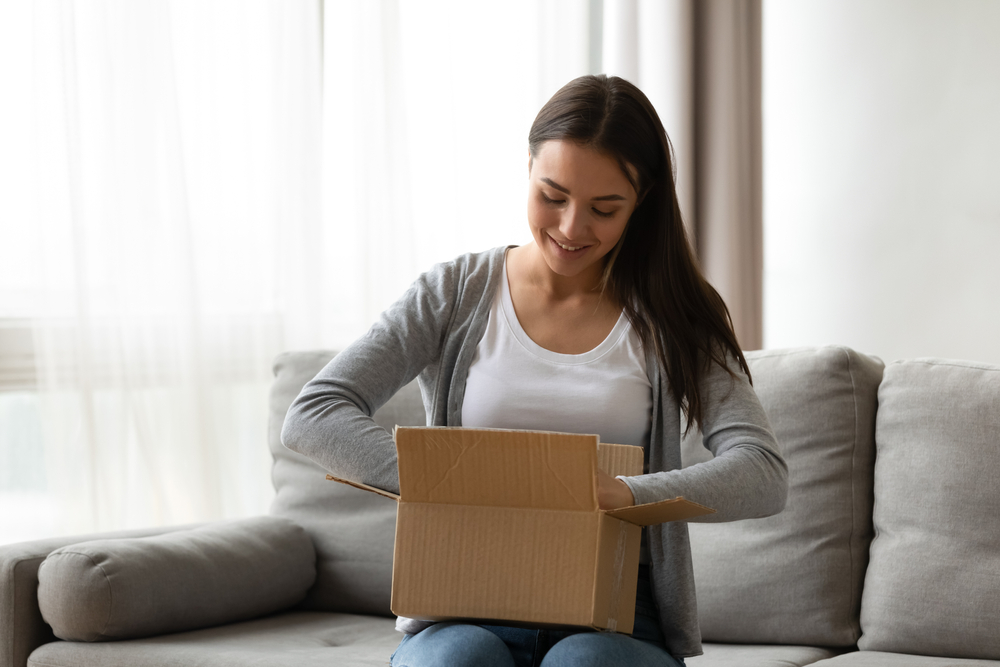A woman sits on a couch while taking an item out of a cardboard box
