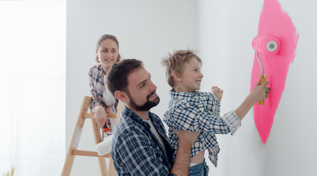 A family paints a wall, a father holding up a child to help