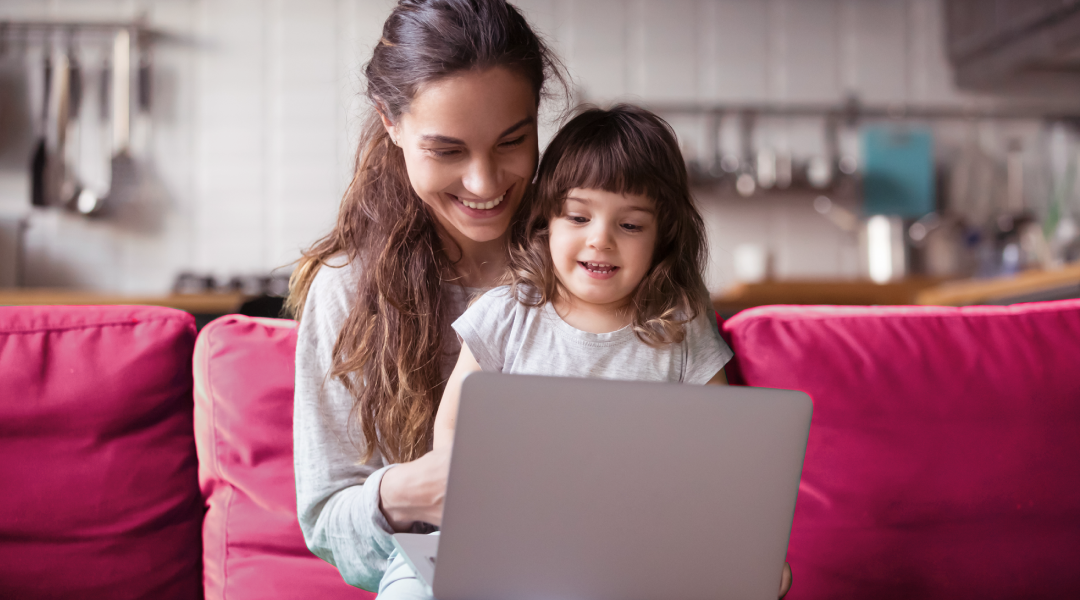 A woman sits with a child on her lap. The child has a laptop on their lap and both people are looking at the laptop.