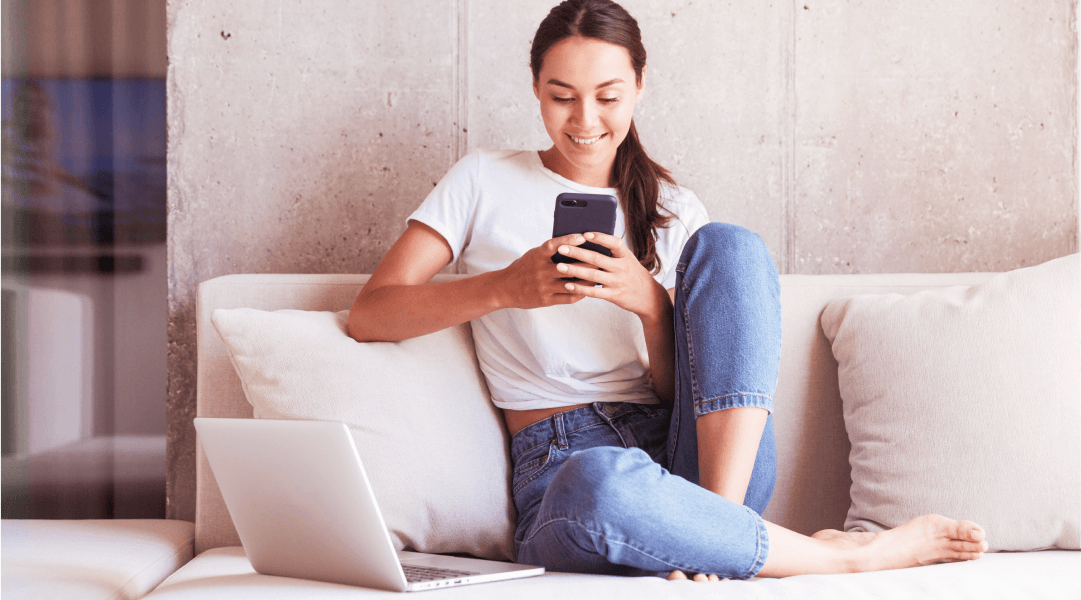 A woman sits on a couch smiling at the smartphone in her hand.