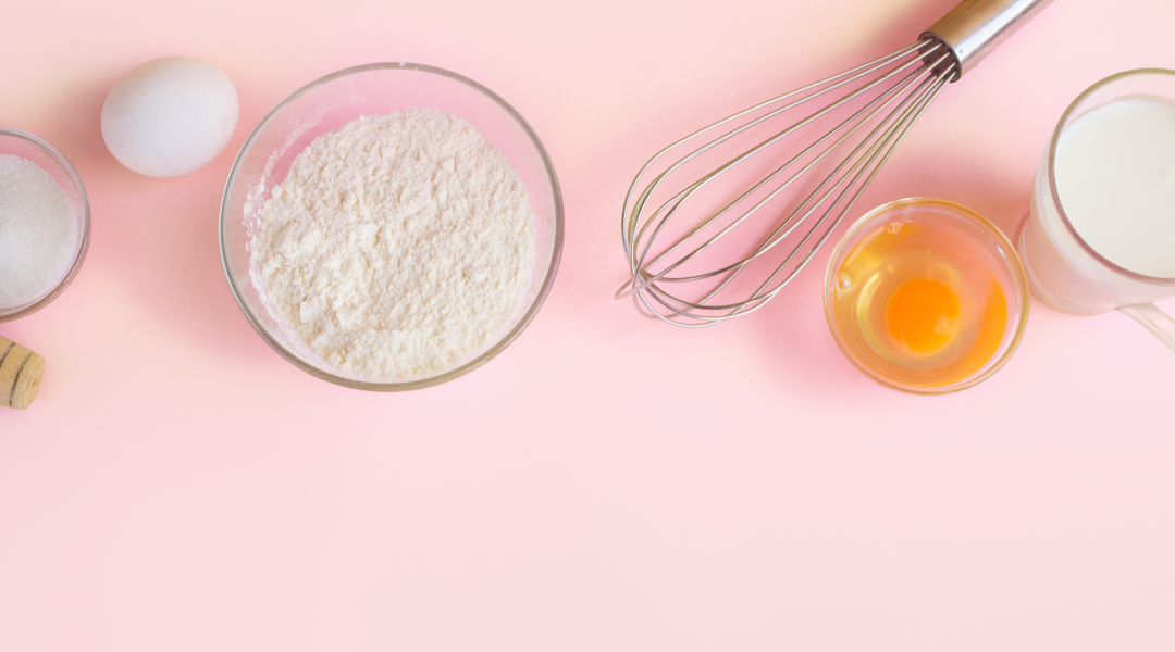 A pink table with a clear bowl of flour, a whisk, and a cracked egg in a bowl