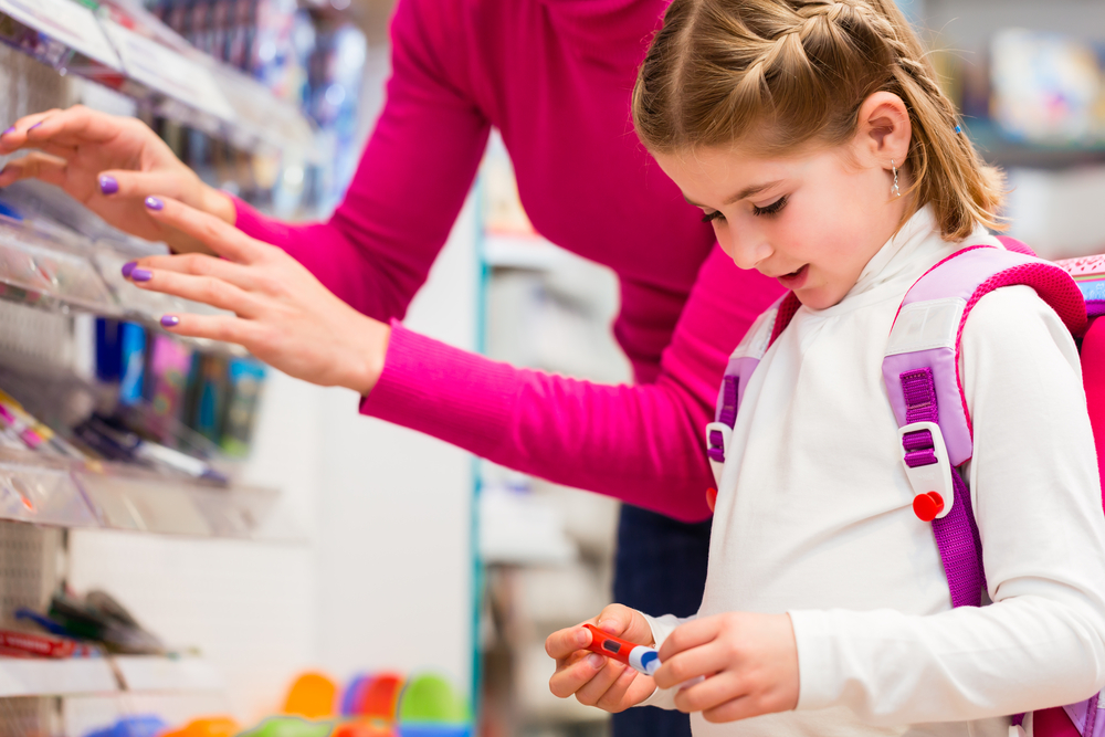 A child holds a school supplies item in a grocery store isle, smiling. Parent is in the background, reaching for another item on the shelf.