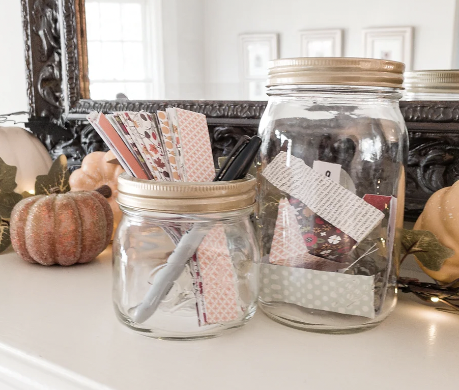 clear glass Ball jars sit on a mantle surrounded by lights and pumpkins. One jar has colorful strips of paper and a Sharpie marker waiting to fill out for gratitudes
