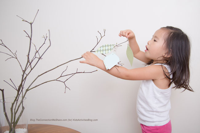 A young child adds paper ornament with gratitudes written on them to a little twig tree in a big clear vase.