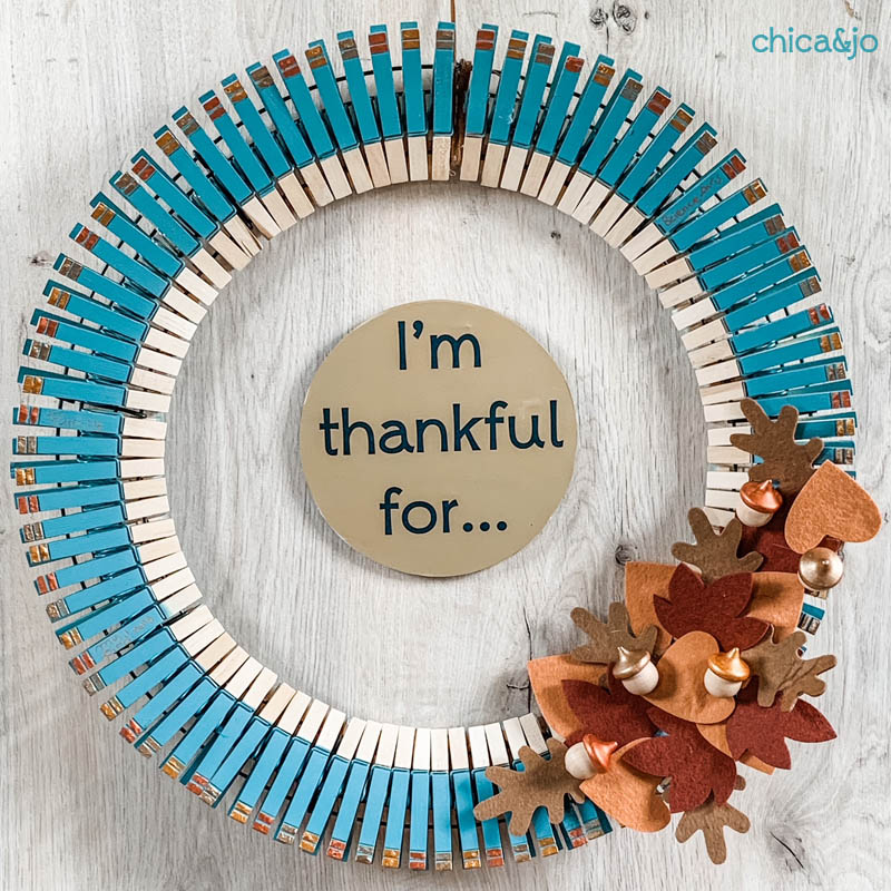 A blue and brown clothespin wreath decorated with orange and brown felt leaves make a Gratitude wreath.